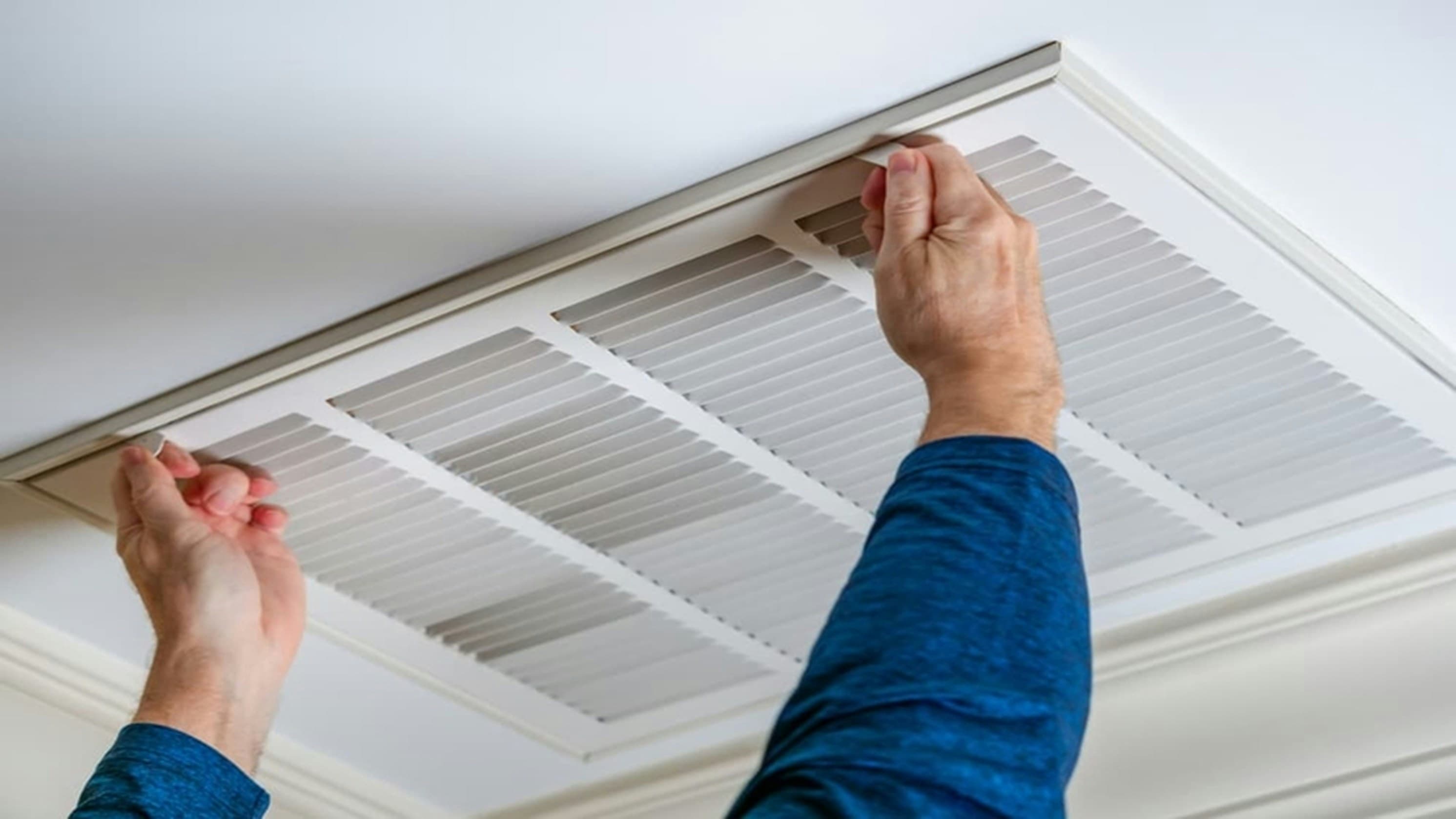 Homeowner removing a dirty gray HVAC air filter from a return air vent in a hallway ceiling