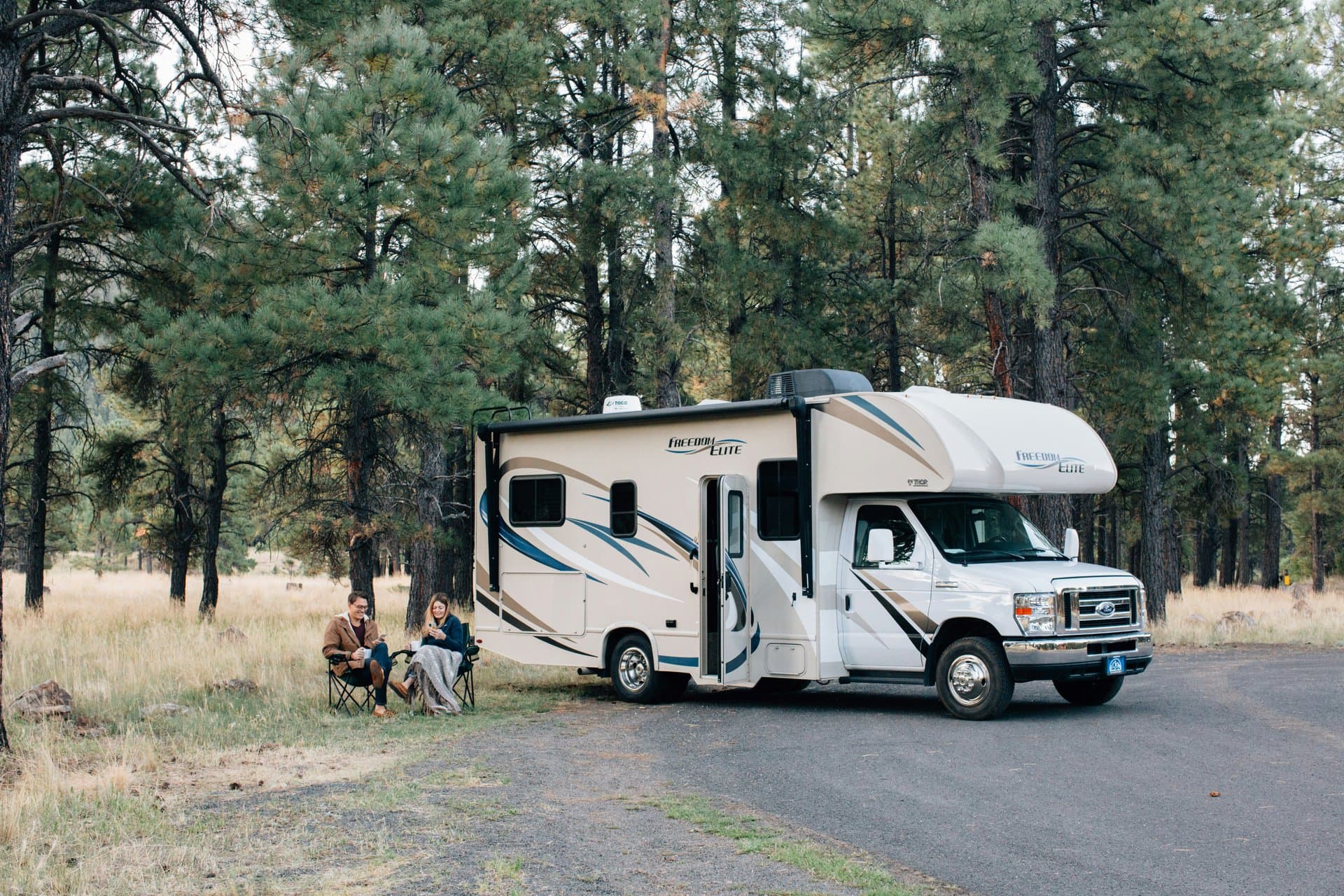 A motorhome parked in a residential driveway on a sunny morning while an owner walks around it checking the tires before a road trip