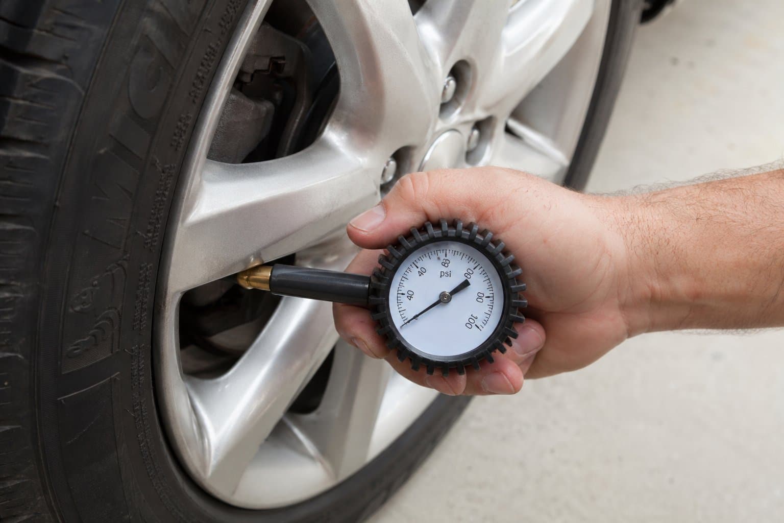 Driver kneeling beside a car tire in a residential driveway, checking pressure with a handheld gauge on a bright spring morning